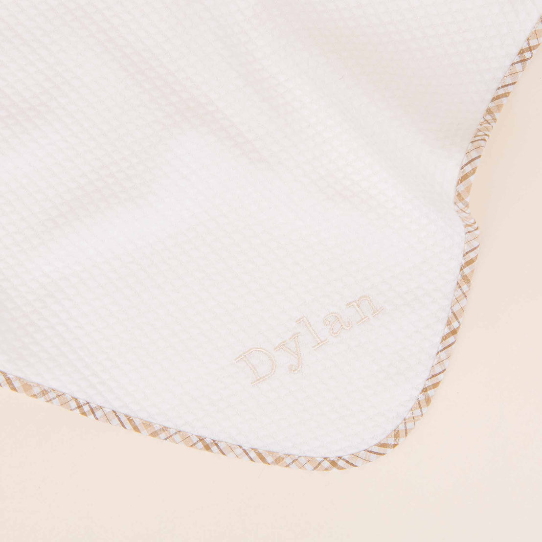 A close-up of a white baby blanket embroidered in a light tan thread. The blanket has a textured diamond pattern and a tan stitched border, perfect for a newborn's coming home.