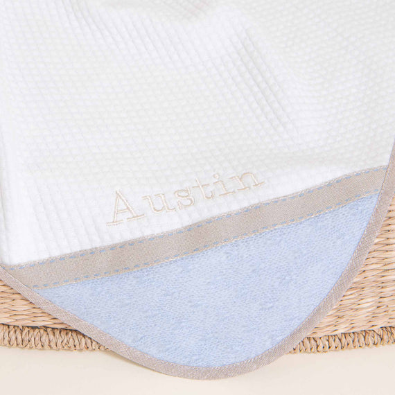A close-up of a white and blue Austin Personalized Blanket with the name "Austin" embroidered in a delicate script, resting in a woven basket.