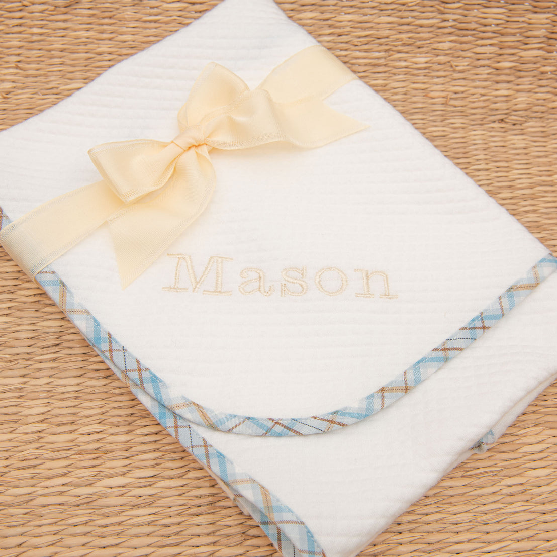 A Mason Newborn Personalized Blanket with the name "Mason" embroidered in gold for a christening, wrapped and decorated with a yellow ribbon bow. The blanket is lined with blue checkered trim, displayed on a vintage wicker background.