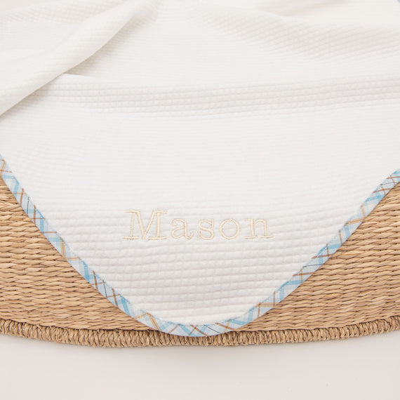 Close-up of a Mason Personalized Blanket with the name "Mason" embroidered in gold on white fabric, edged with a blue patterned trim, arranged over a woven basket, perfect for a coming home or baptism.