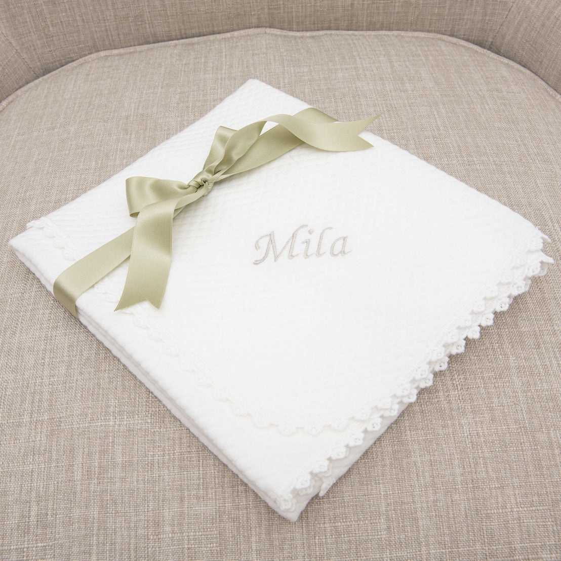 The Mila personalized blanket with the name "Mila" embroidered in silver thread, designed as an heirloom for a newborn, adorned with a sage green ribbon, neatly placed on a textured beige fabric chair.
