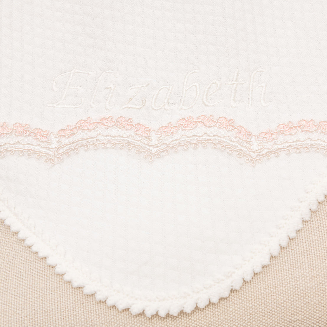 Close-up of a personalized white heirloom Blanket with the embroidered name "Elizabeth" above a heart-shaped lace pattern with pink detailing, ideal for a christening.