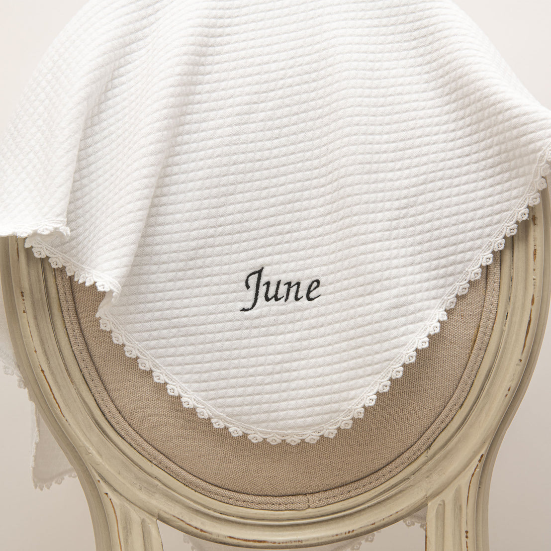 A white textured June Personalized Blanket with the name "June" embroidered in black, draped over an elegant vintage cream-colored chair with intricate details.