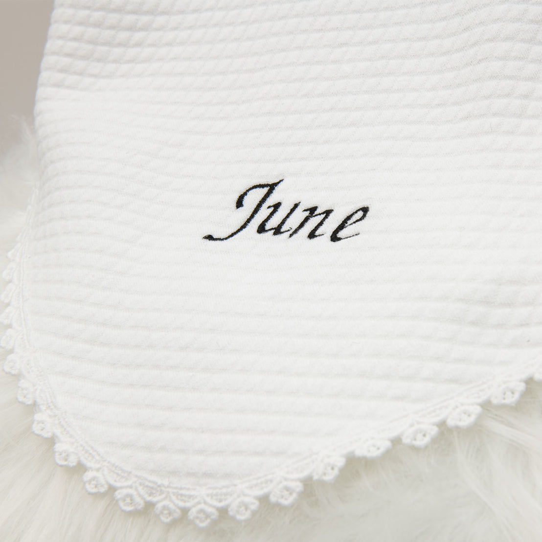 Close-up of a white textured cotton fabric with the name "June Personalized Blanket" embroidered in elegant black script, edged with delicate lace detailing, perfect as a personalized gift.