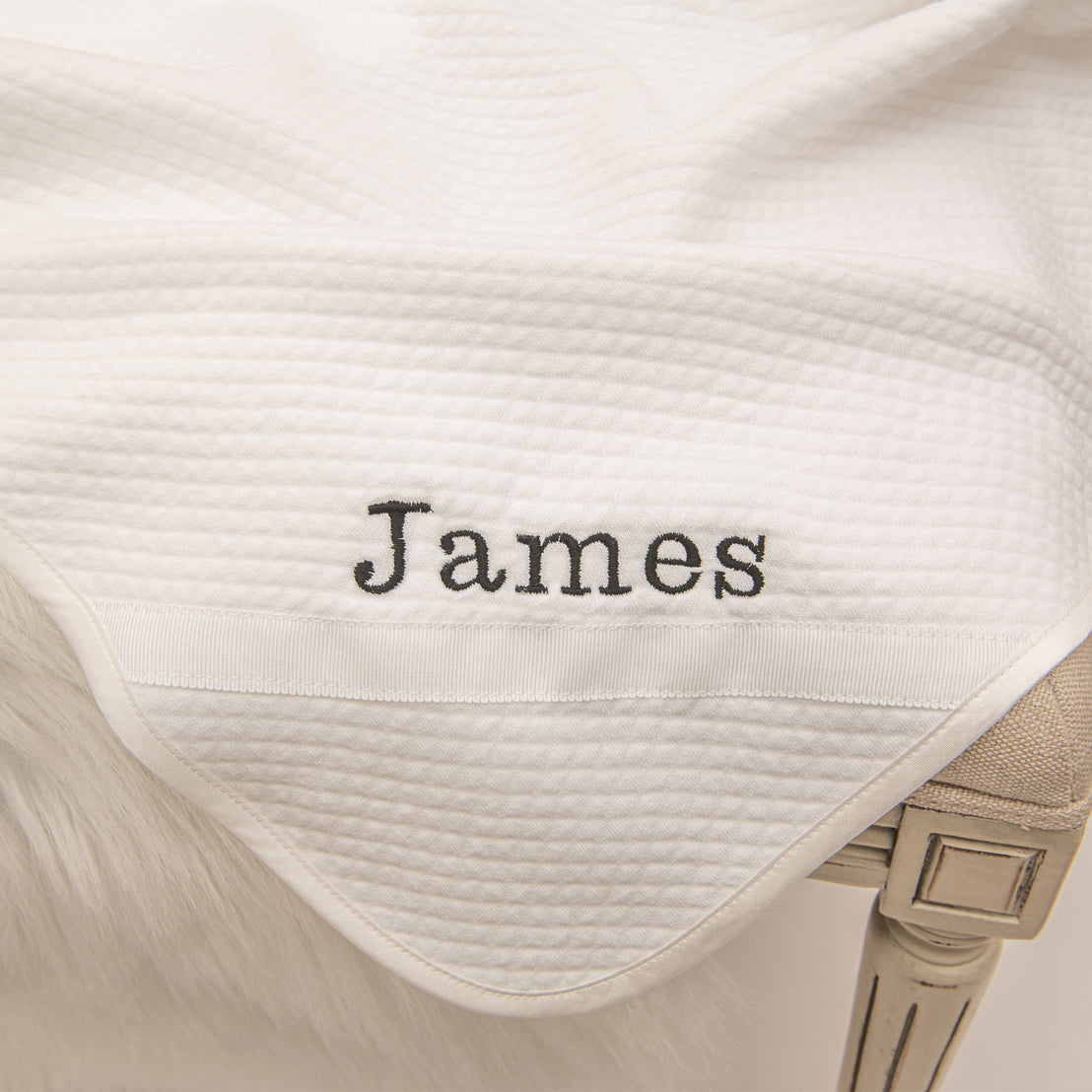 Flat lay photo of the James Personalized Blanket. It is made from 100% white textured cotton with white grosgrain ribbon, silk ribbon, and the name "James" embroidered on the corner.