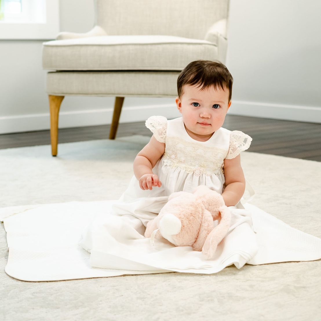 A baby with dark hair sits on the Jessica Personalized Blanket, holding a plush pink bunny, dressed in the Jessica Linen Gown, with a light grey armchair and white walls in the background.