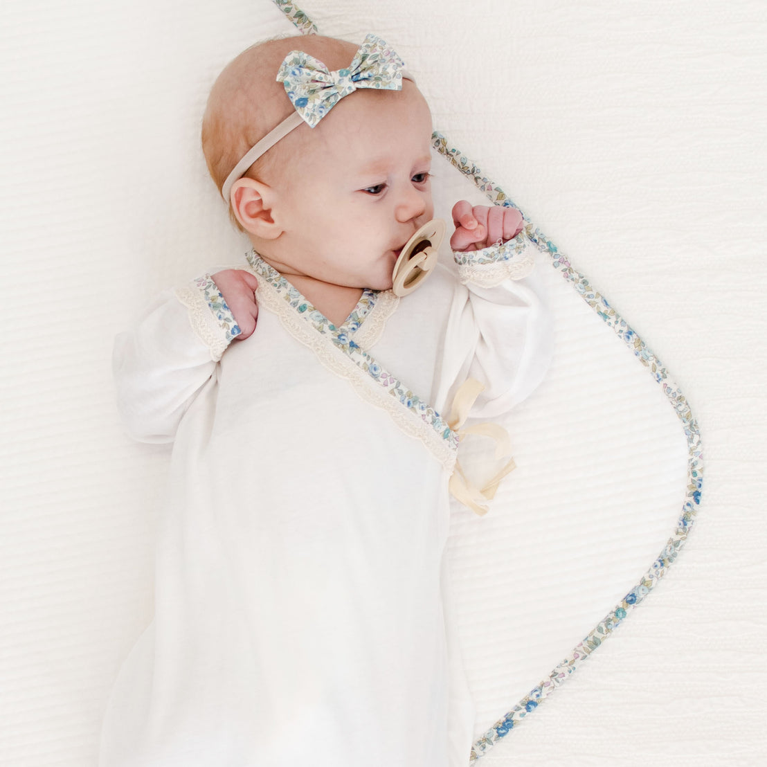 A baby with a light blue bow headband and a pacifier lies on an upscale white background, encircled by a Petite Fleur Personalized Blanket.