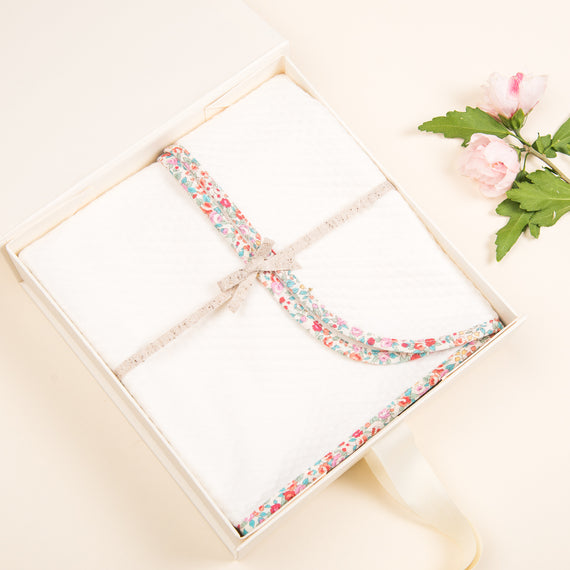 An elegant upscale Petite Fleur Personalized Blanket with a floral fabric ribbon, placed next to a pink flower on a soft beige background.