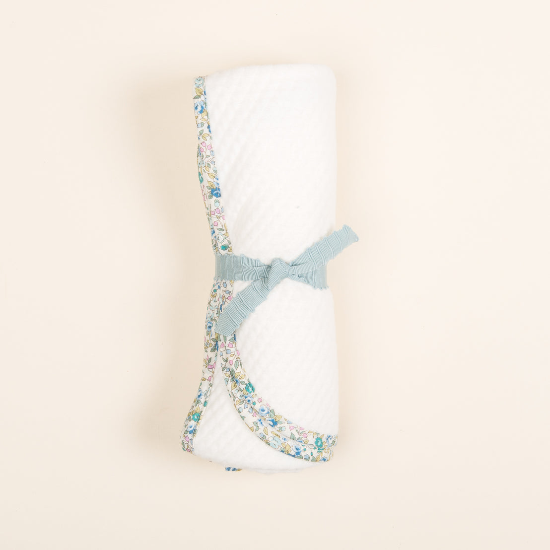 A rolled Petite Fleur Personalized Blanket tied with a light blue ribbon and accented with a floral fabric trim against a pale beige background, featuring Vintage-inspired details.