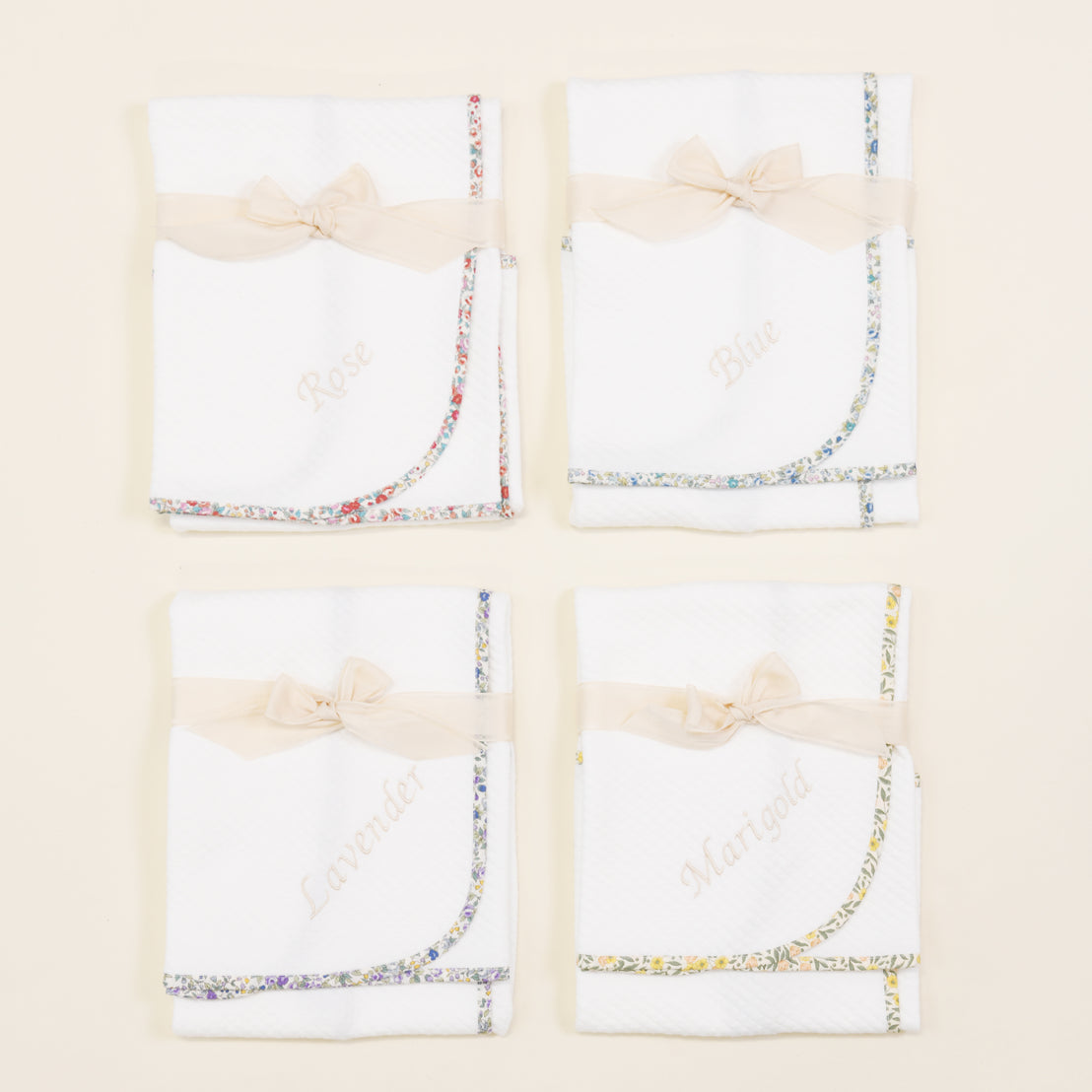 Four white Petite Fleur Personalized Blanket fabric swatches decorated with floral trim and satin bows, labeled rose, blue, lavender, and marigold, displayed on a light background.