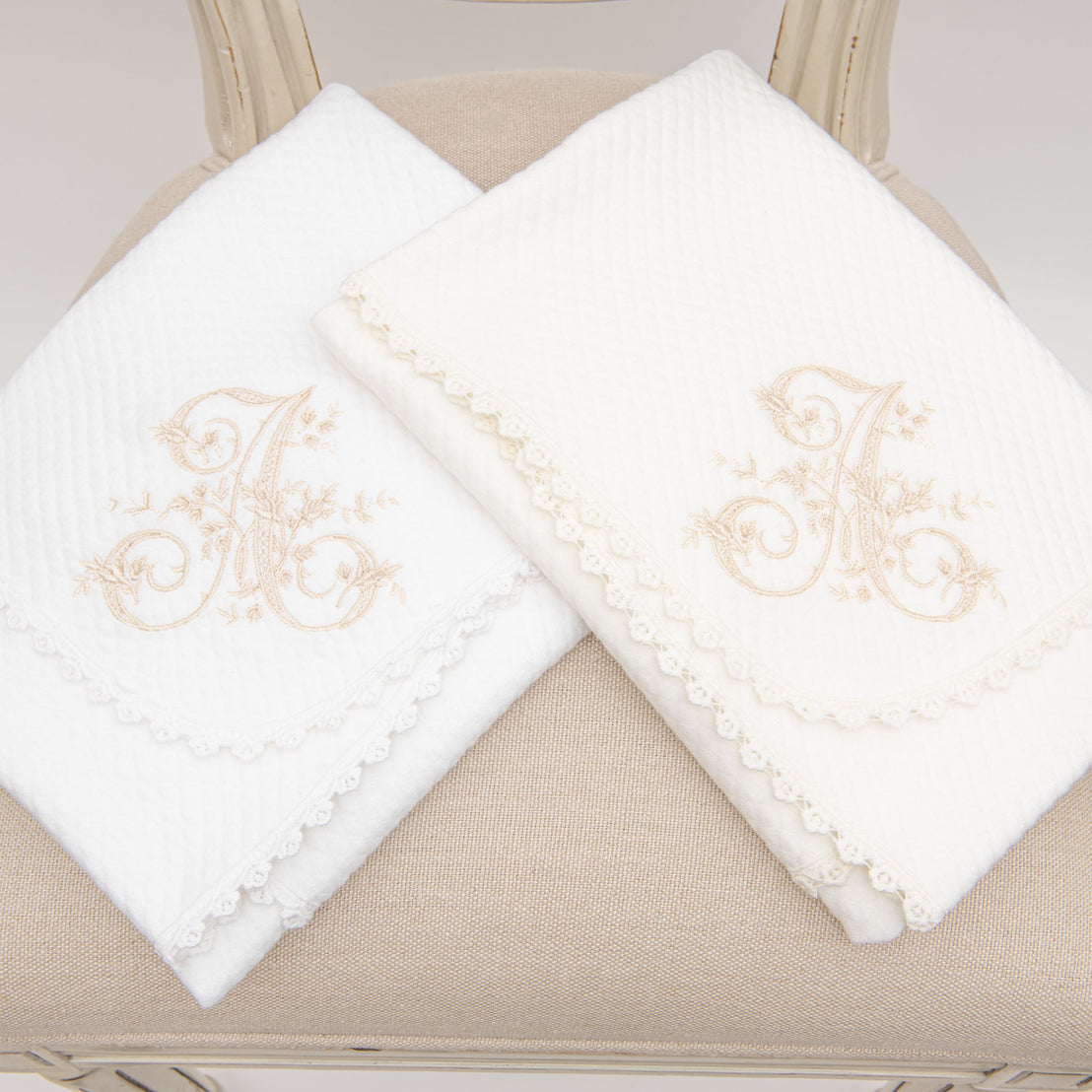 Two white embroidered baby blankets with ornate gold initial "A" designs, displayed on a beige chair with intricately detailed lace edges. These blankets are made from 100% quilted cotton.