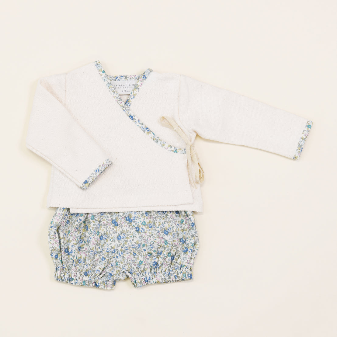 An upscale, vintage-inspired Petite Fleur Wrap Top & Bloomers set laid flat on a light beige background. The top is white with floral trim, and the shorts feature a blue floral print.