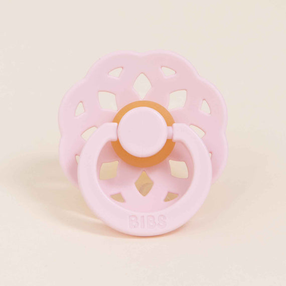 A Bibs Lace Pacifier in Blossom with a circular, scalloped shield and an orange button handle, designed in Denmark, set against a light beige background.