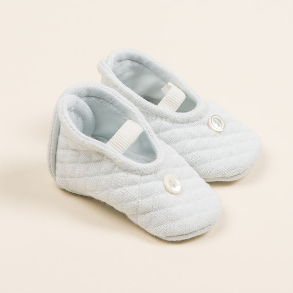 A pair of upscale, light blue, Aiden Quilted Booties with a velcro strap and a button detail, set against a soft beige background.