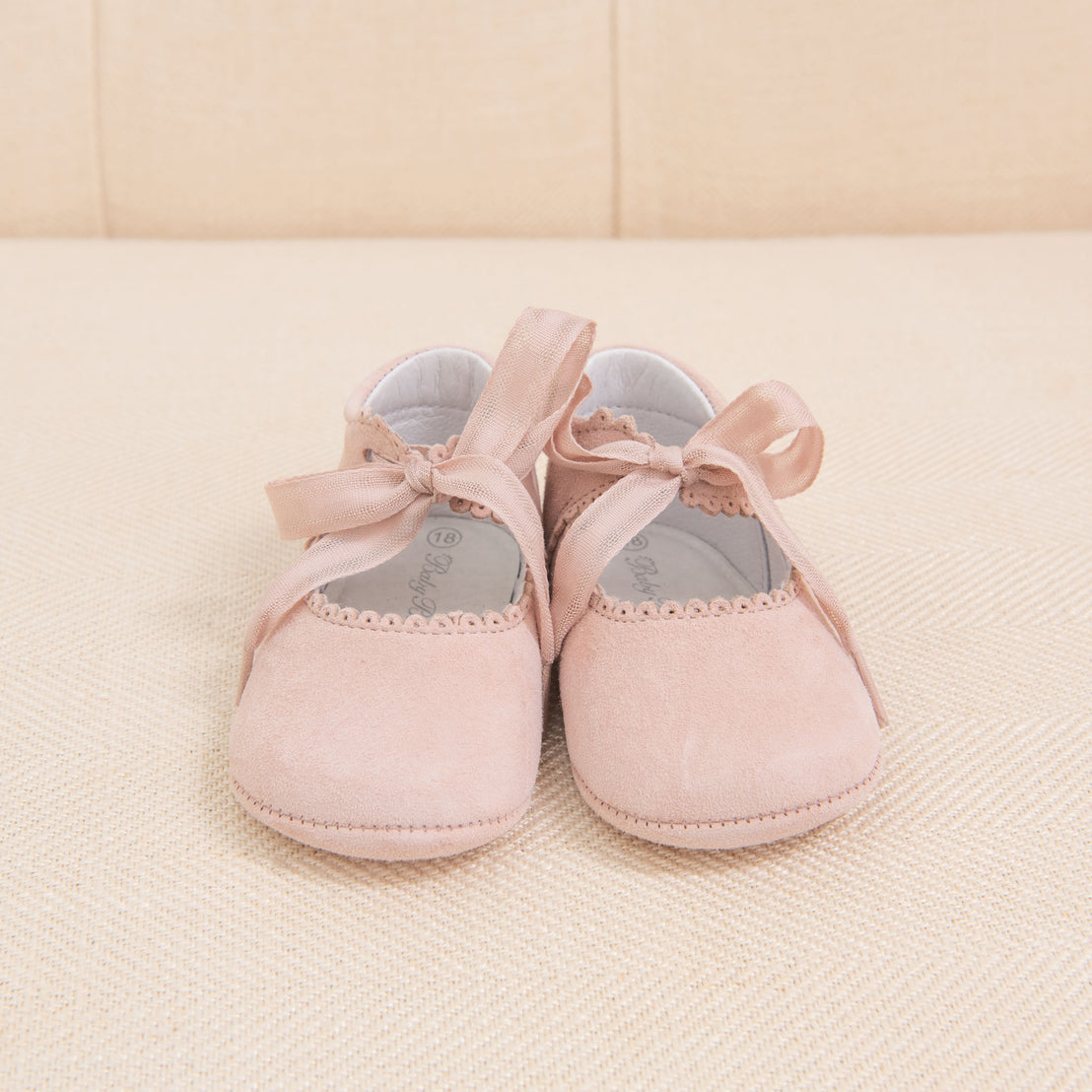 A pair of small pink Girls Suede Tie Mary Janes with ribbon ties, placed on a beige textured fabric background.