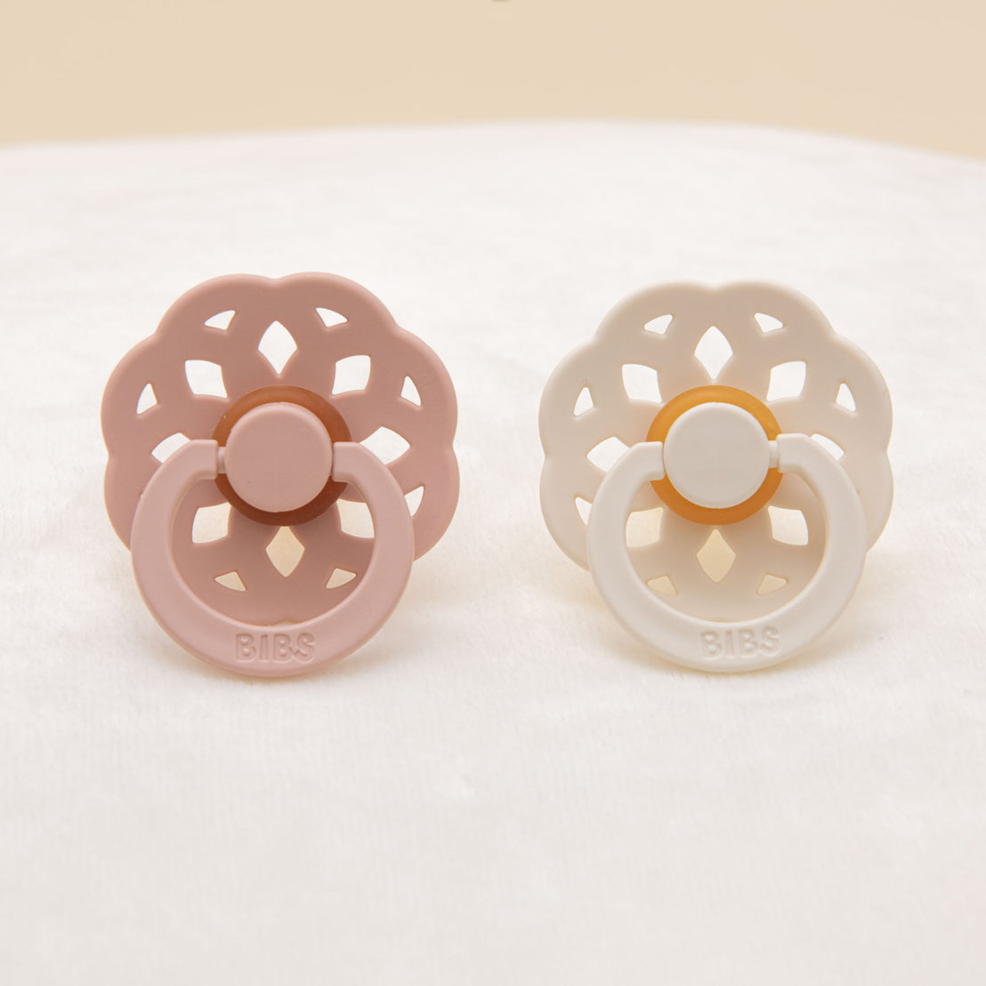 Two Isabella Pacifiers, one in Ivory & the other in Blush, are placed side by side on a soft beige fabric.