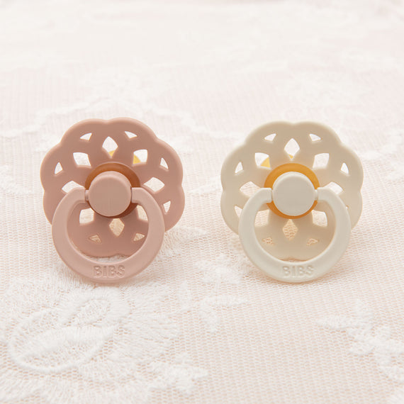 Two Isabella Pacifier Sets, one pink and one cream, styled with a vintage floral design around the shield, resting on a textured white fabric.