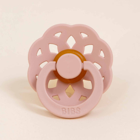 A soft pink Bibs Lace Pacifier 2 Pack | Blush with a round handle and large circular vent holes, featuring the brand name "bibs" centered on a beige button. The background is plain and light-colored. 🍼