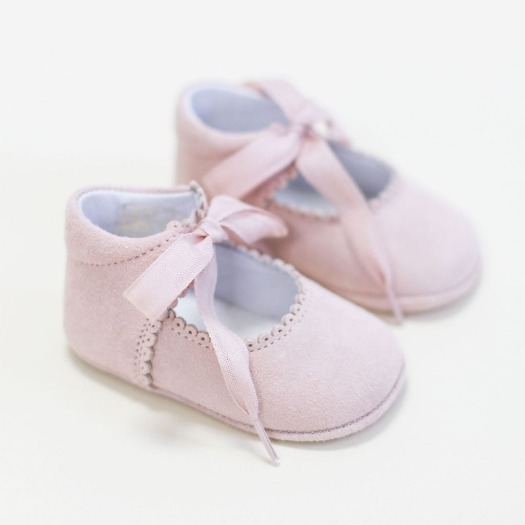 A pair of Blush Suede Tie Mary Janes with delicate ribbon laces and detailed stitching against a light, neutral background.
