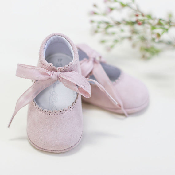 A pair of Blush Suede Tie Mary Janes with delicate bow ties, partially adorned by a subtle touch of greenery in the top right corner.