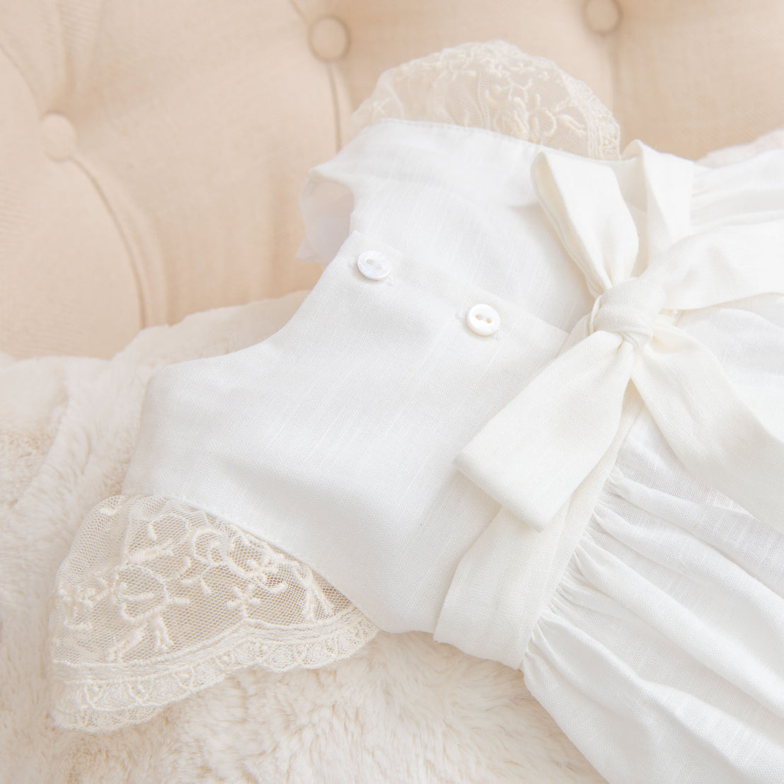 Close-up of the back of the Jessica Linen Gown showcasing the backside pearl buttons, white bow, and lace sleeves, placed on a soft beige cushion.