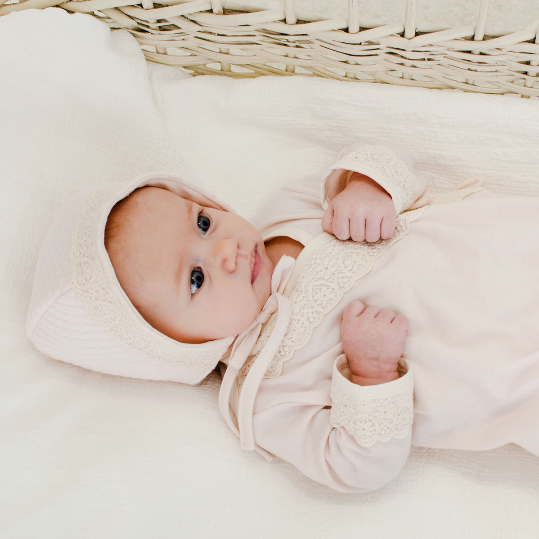 A newborn baby dressed the Evelyn Knot Gown outfit with lace details and Evelyn Quilt Bonnet lies in a woven basket, looking curiously upward..