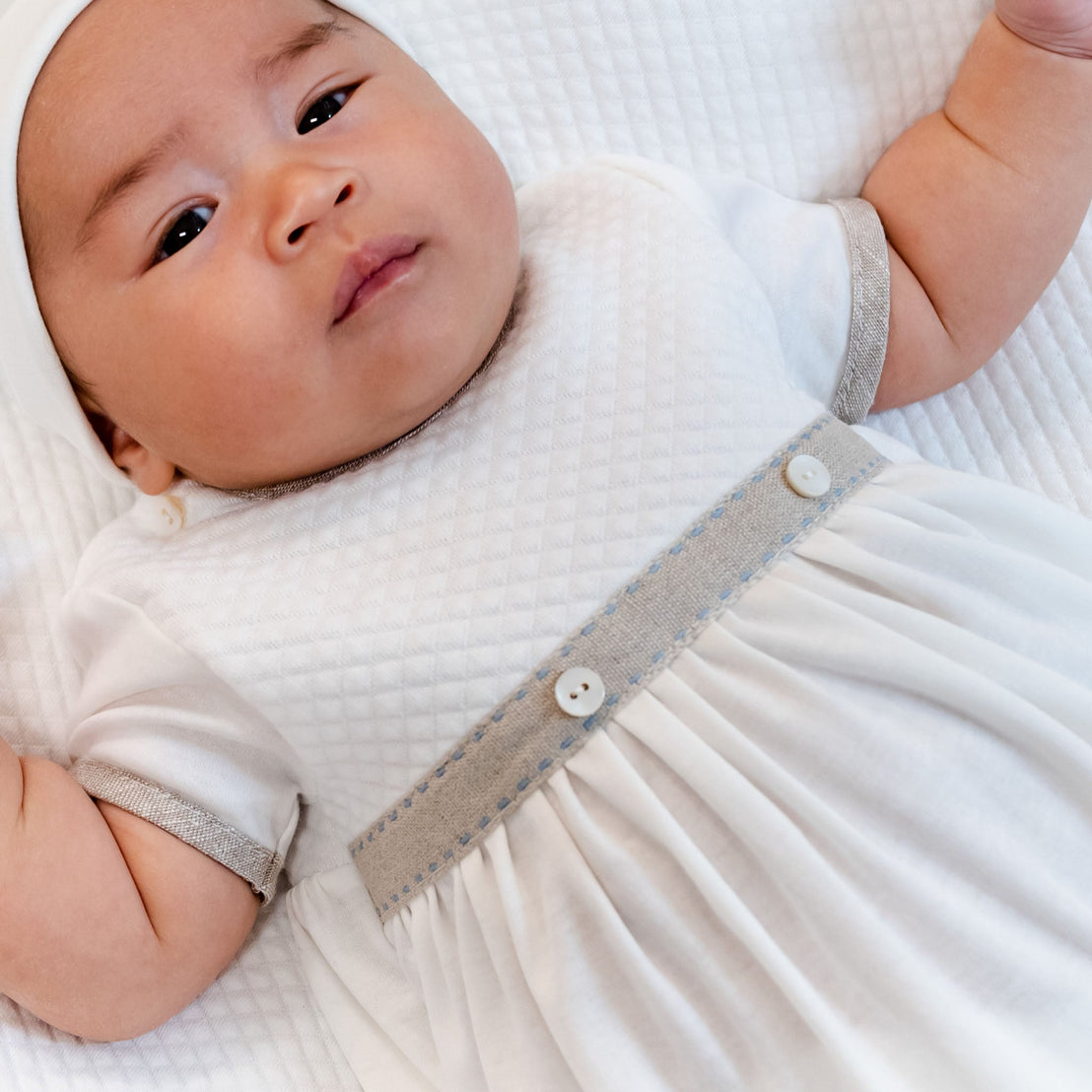 A close-up of a baby lying on a white blanket, wearing the Austin Layette & Hat, looking upwards with a calm expression.