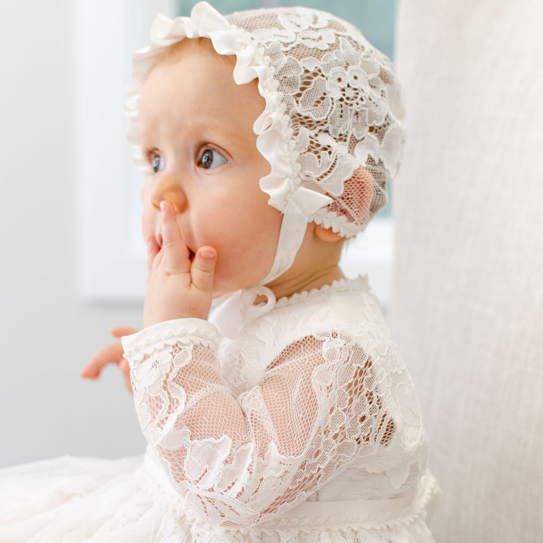 Baby girl with finger in mouth wearing a long-sleeved baptism gown and also a lace baptism bonnet.