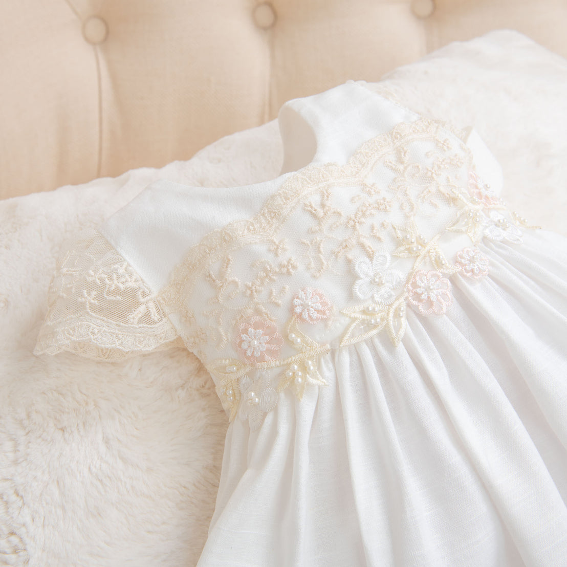 An elegant Jessica Linen Gown with delicate lace and floral embroidery detail, laid out on a soft, cream-colored cushioned background.