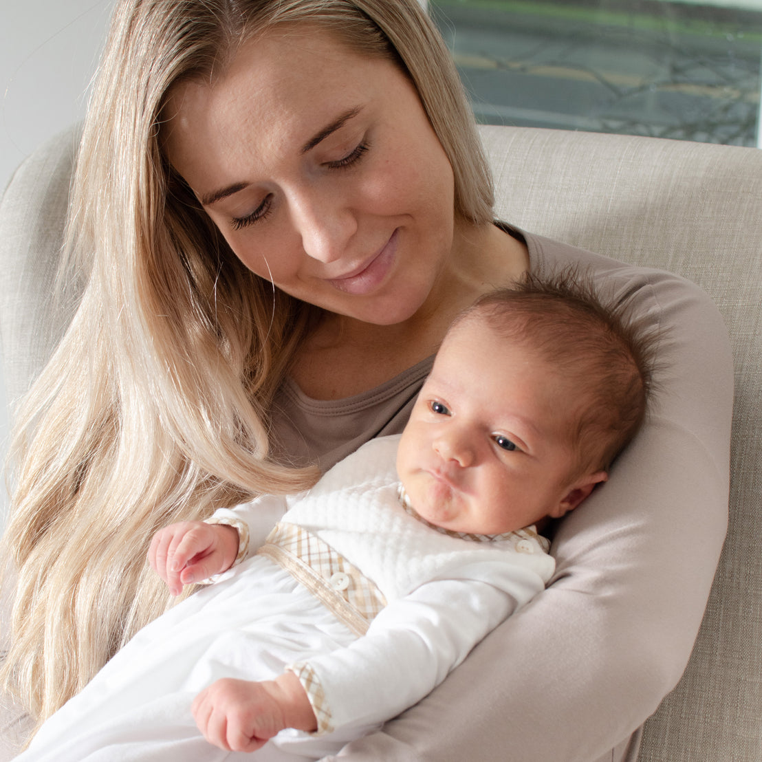 A tender moment between a woman and a newborn baby. The woman, smiling gently, cradles the wide-eyed baby in her arms while sitting in a cozy chair. The newborn is snug in a Dylan Layette.