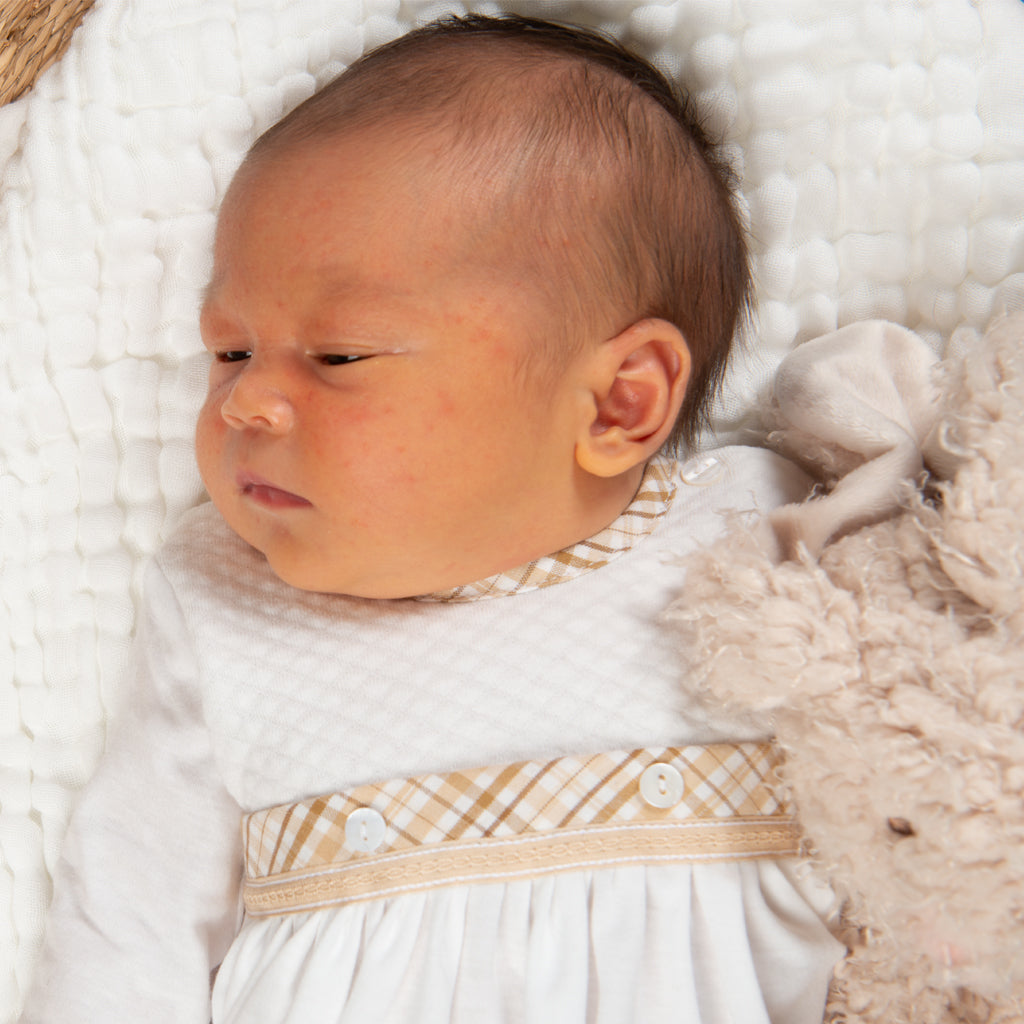 A newborn baby with light brown hair, lying on a white blanket, wearing a Dylan Layette outfit with a beige patterned trim, next to a soft, fluffy beige blanket.