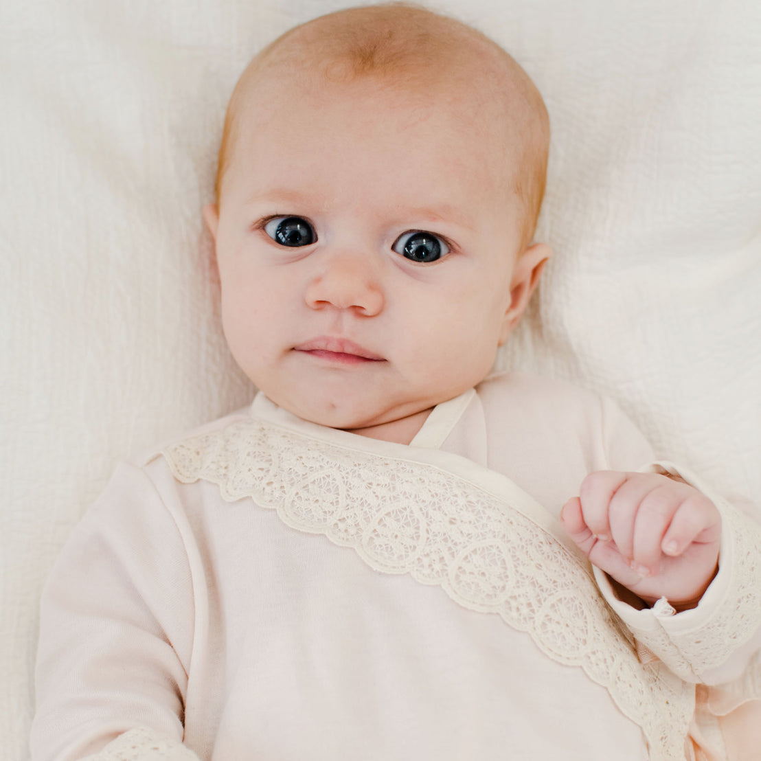 A newborn with wide blue eyes and light hair, wears the Evelyn Knot Gown with lace detailing, and lies on a soft white background.