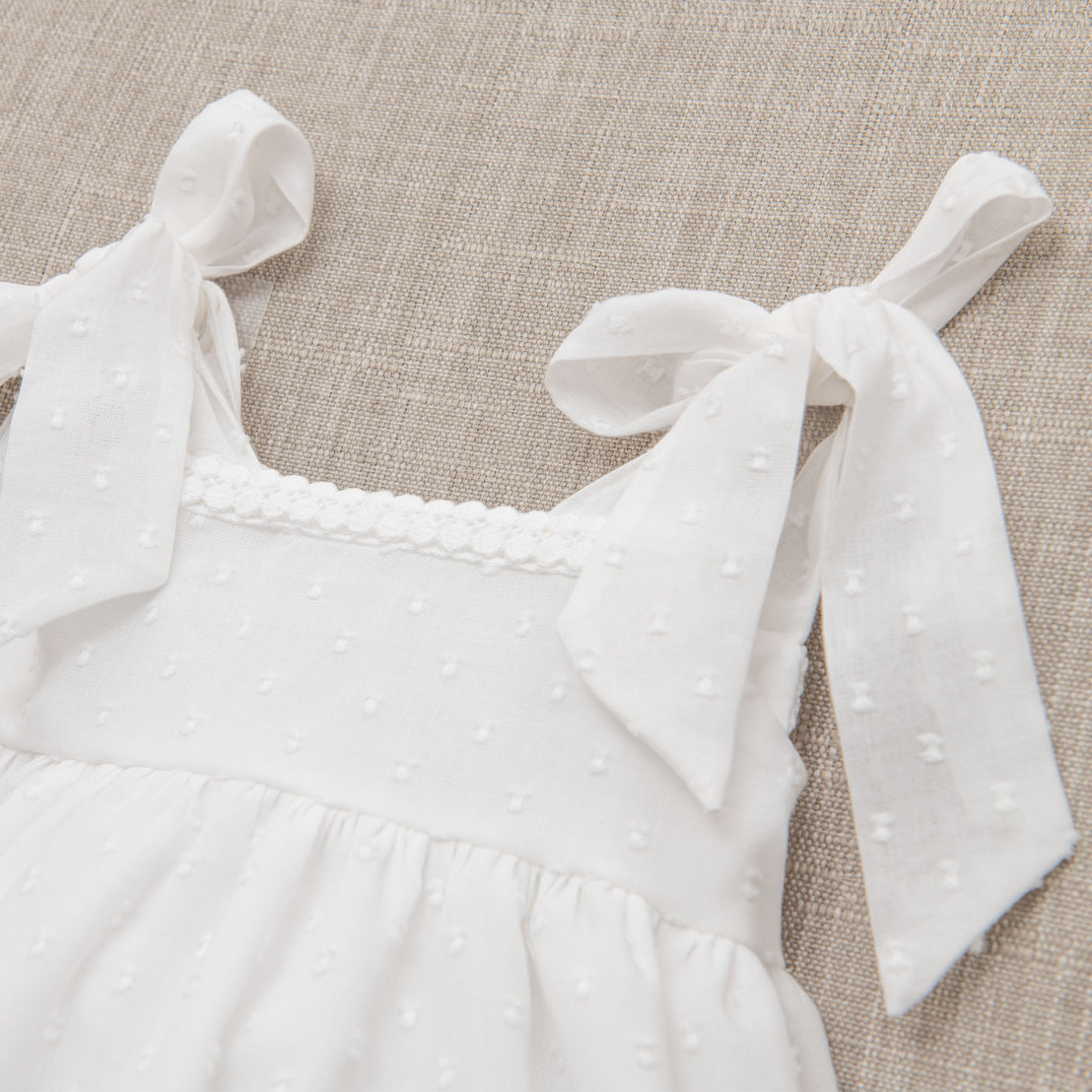 Close-up of Mila Cotton Gown made from Montreux Swiss Dot cotton, a textured dot pattern fabric, featuring a detailed neckline and ribbon-tied shoulder straps, displayed on a beige fabric background.