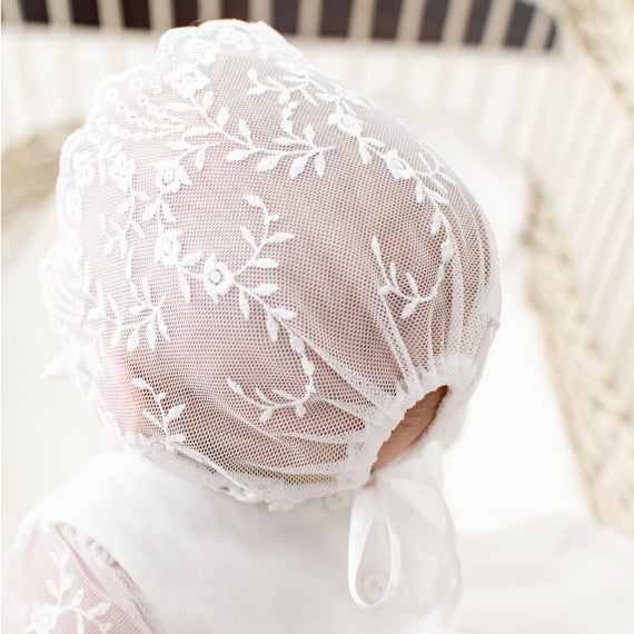 A newborn wearing an Isla Lace Bonnet with floral embroidery, viewed from the back, inside a crib.