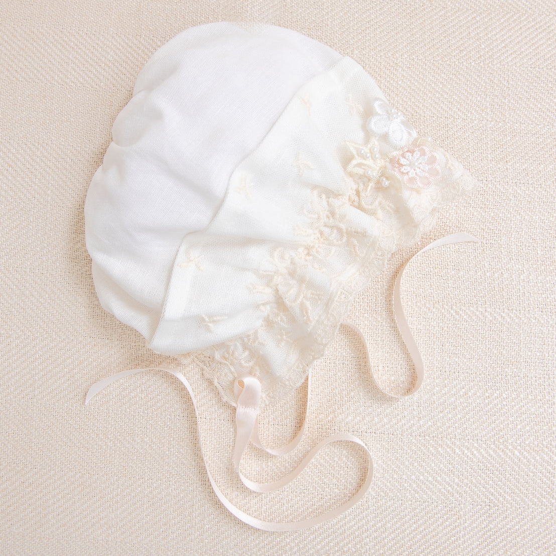 The Jessica Linen Bonnet, a delicate white bonnet with champagne lace detailing and thin ribbon ties, laid out on a textured cream background.