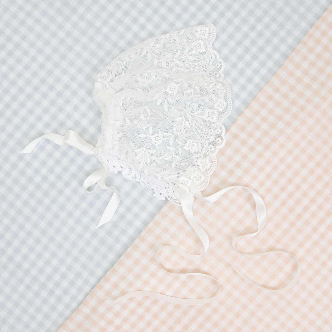 A delicate Isla Lace Bonnet with satin ribbon ties, ideal for a newborn, displayed on a pastel blue and peach checkered background.