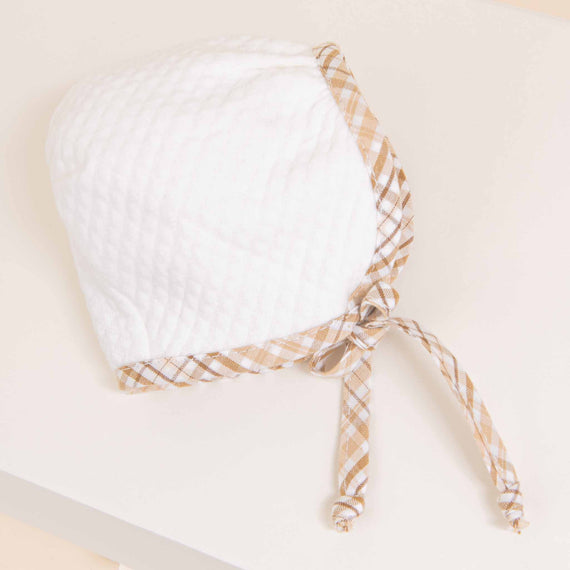 A white textured Dylan Quilted Newborn Bonnet with beige checked trim and straps, perfect for a newborn's coming home from hospital outfit, displayed on a light beige background.