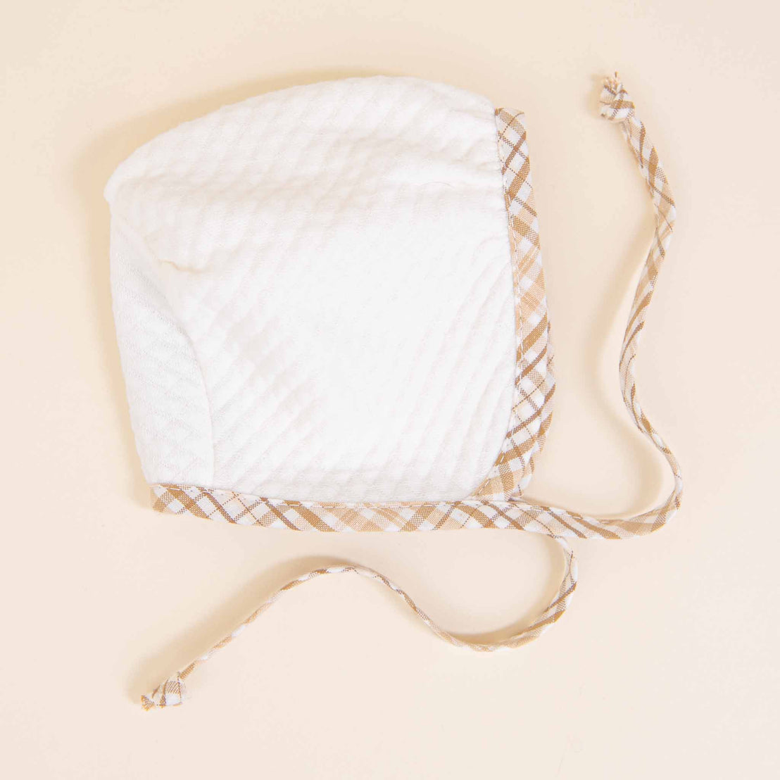 A white Dylan Quilted Newborn Bonnet with tan trim and ties lies flat on a soft beige background. The bonnet is quilted and has a subtle pattern, giving it a gentle texture.