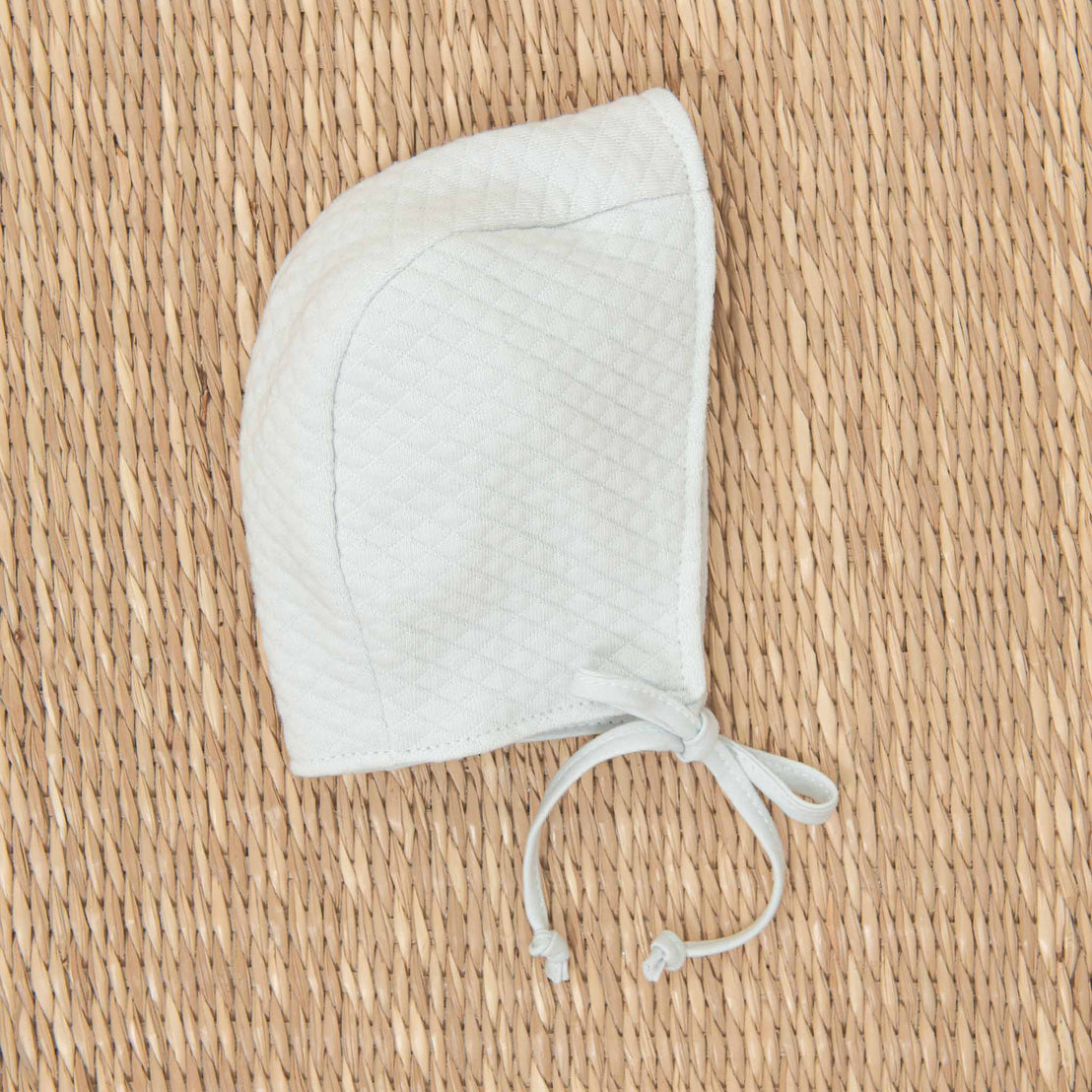 A white Aiden Quilt Bonnet with tie strings, perfect for coming home from the hospital, placed neatly on a straw-textured background.
