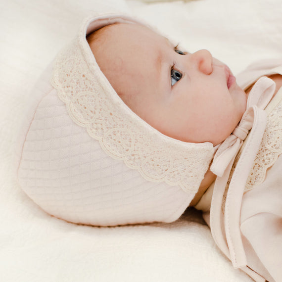 A newborn baby wearing an Evelyn Quilt Bonnet adorned with lace, lying on a white soft surface, gazing upward with clear blue eyes.