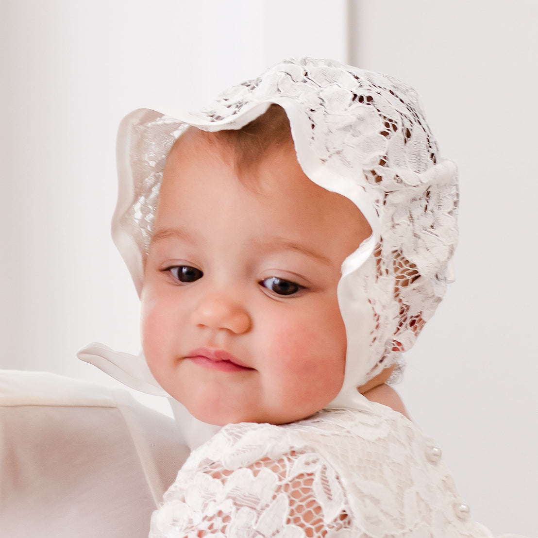 A close-up of a baby wearing a Rose Lace Bonnet and dress with a thoughtful expression, set against a soft white background.