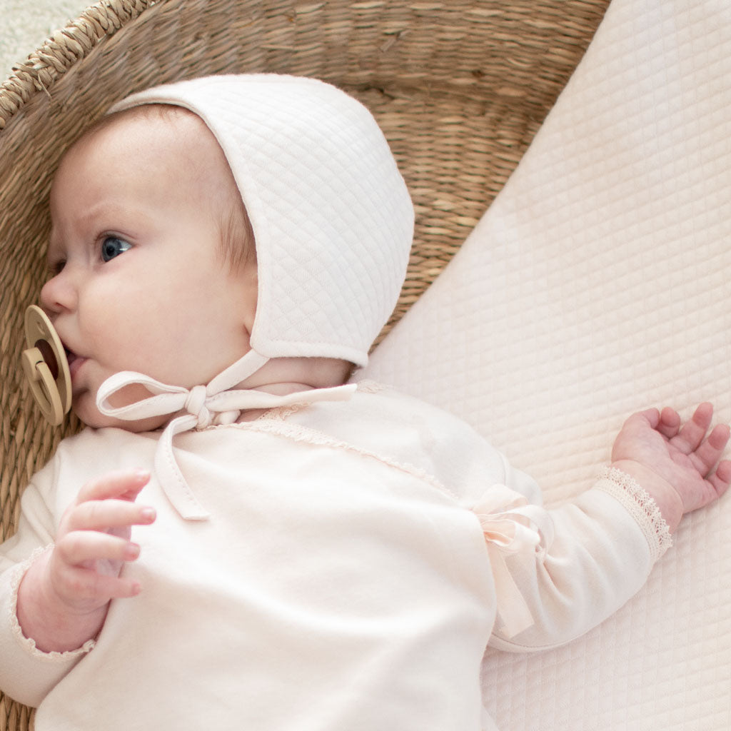 A baby wearing the Ava Quilt Bonnet and Ava Newborn Layette lies in a basket with a blush pink quilted blanket underneath, and sucks on a pacifier.