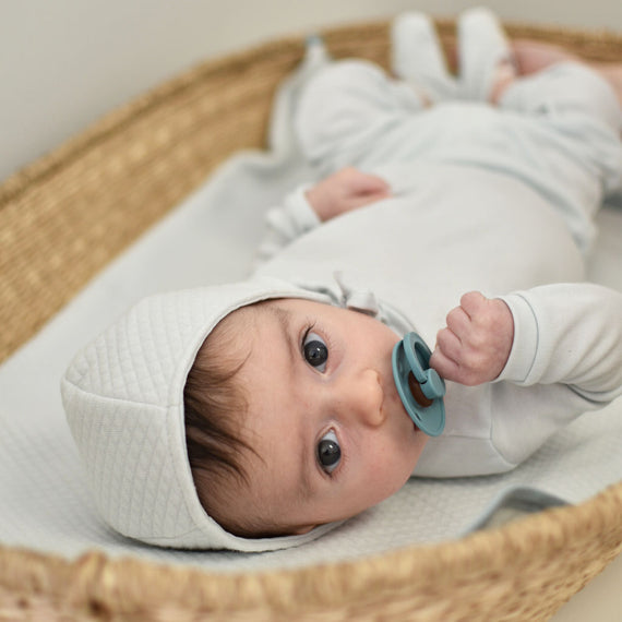 A newborn baby wearing an Aiden Quilt Bonnet and outfit lies in a wicker basket, holding a blue pacifier and gazing upward with wide eyes.