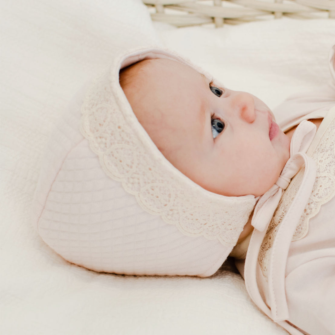 A close-up of a newborn baby wearing the Evelyn Quilt Bonnet, looking up with wide blue eyes, along with the Evelyn Knot Gown..