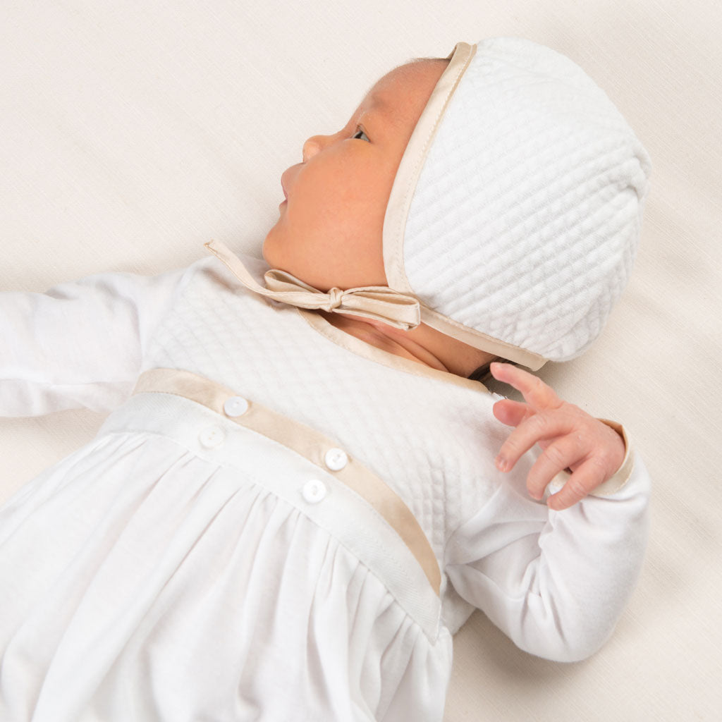 Newborn baby wearing the Liam Quilted Newborn Bonnet made with soft quilted cotton in ivory and trimmed with a champagne colored silk