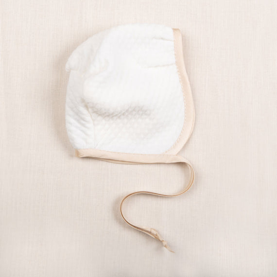Flat lay photo of the Liam Quilted Newborn Bonnet made with soft quilted cotton in ivory and trimmed with a champagne colored silk