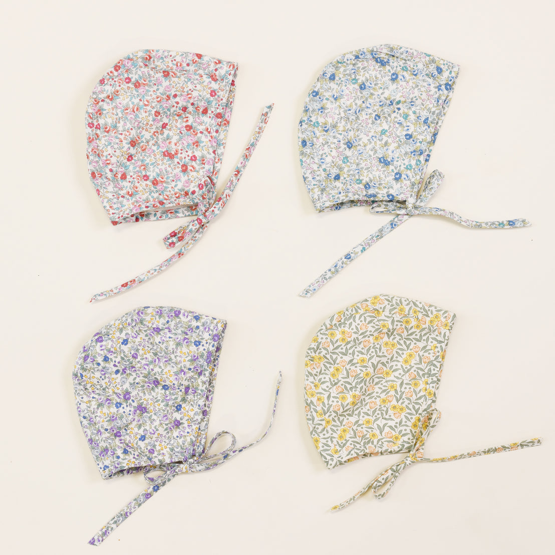 Four upscale, Petite Fleur Bonnets are arranged in a square pattern on a white background, each featuring fabric ties and a unique floral design.