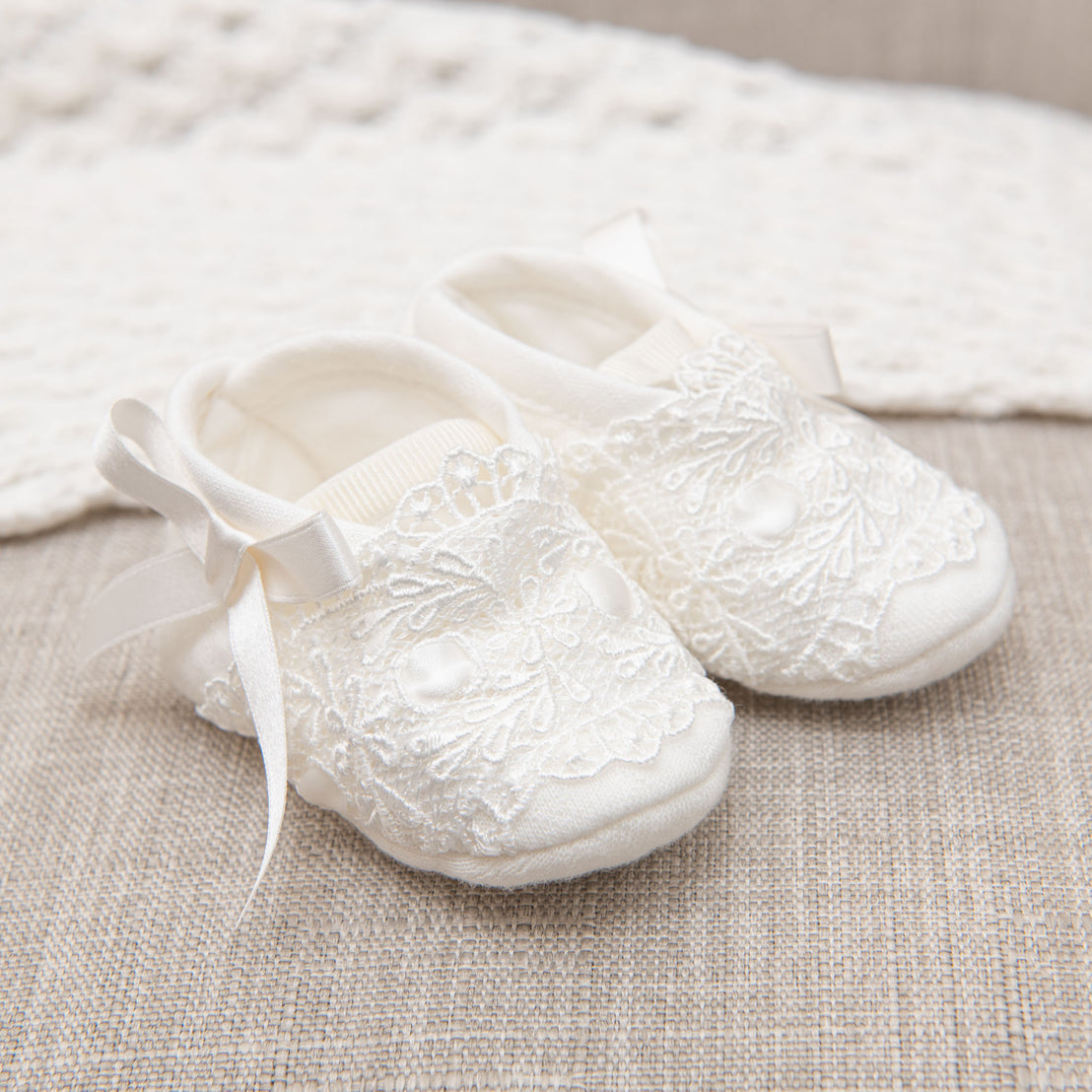 A Madeline Newborn Gift Set with delicate white heirloom baby shoes with lace details and satin ribbons, placed on a soft beige fabric surface. 