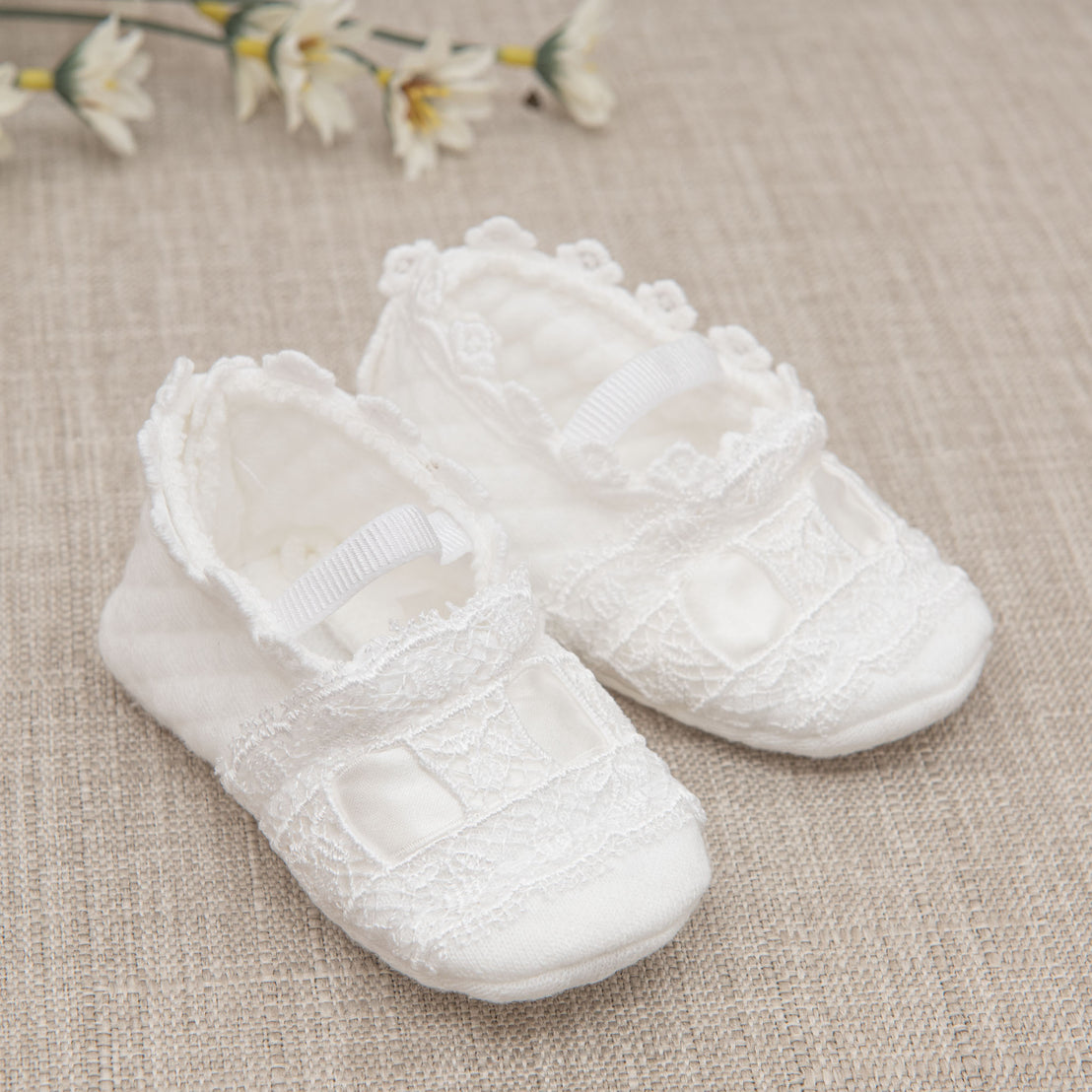 A pair of white lace Mila baby shoes with ribbon detailing, a unique baby gift, sits on a textured beige surface, accompanied by soft-focus white flowers in the background.
