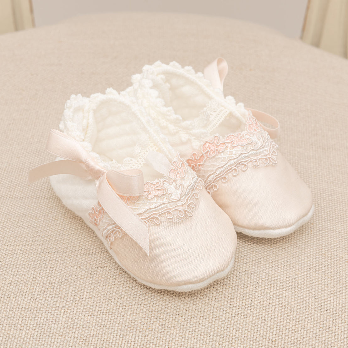 A pair of delicate, pink baby Elizabeth shoes with lace detailing and ribbon ties, neatly placed on a soft beige textured surface, perfect for a christening.
